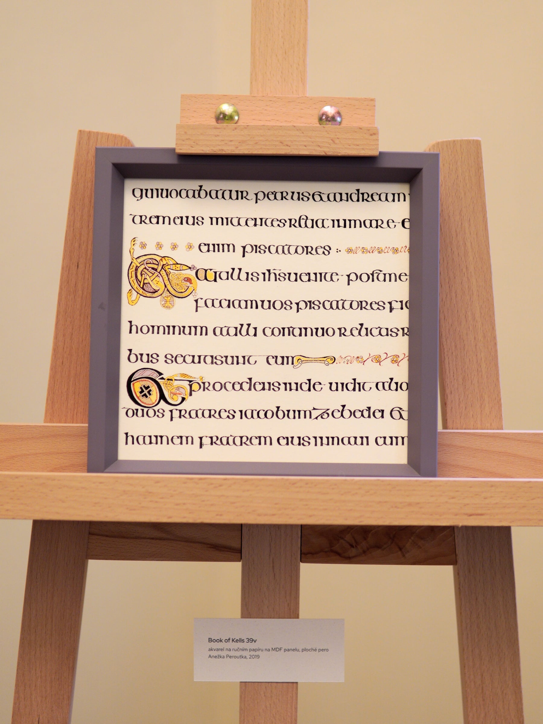 Kopie listu z Book of Kells v rámu na dřevěném stojanu, s popiskem.